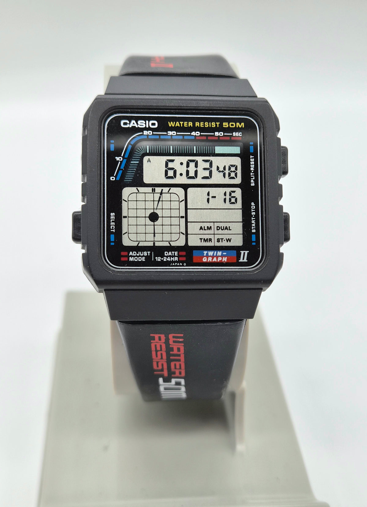 A Casio square analog-digital wristwatch with a black case and strap, featuring a graphic face design with blue, red, and white details, displayed on a light gray stand.