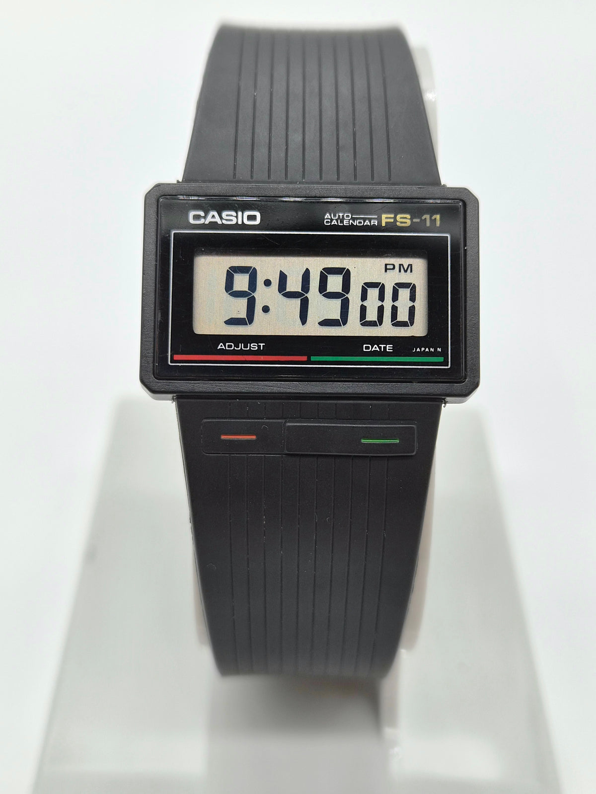 A vintage Casio EX-O-Matic FS-11 digital watch with a black ribbed strap and a rectangular face, displayed on a white stand against a white background.