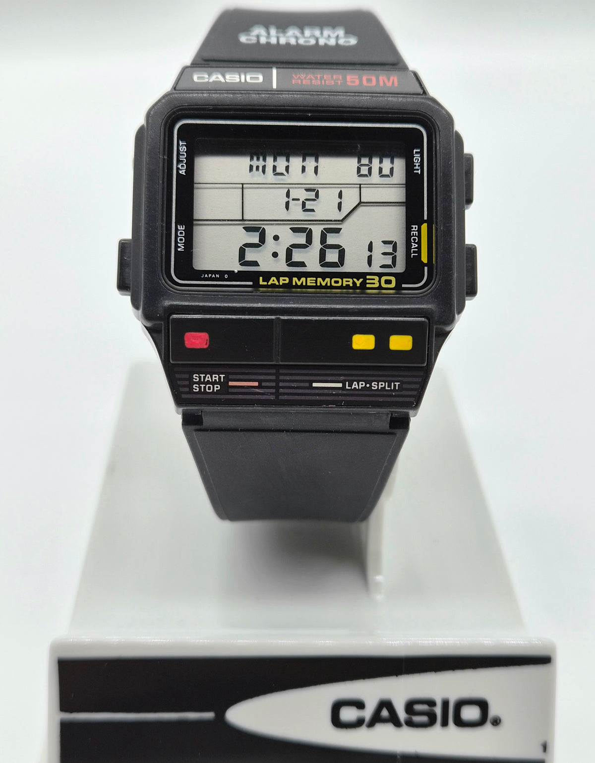 A Casio digital alarm watch with a black strap and case, featuring a digital LCD display and multiple control buttons, shown resting on a white display stand with a Casio-branded base.