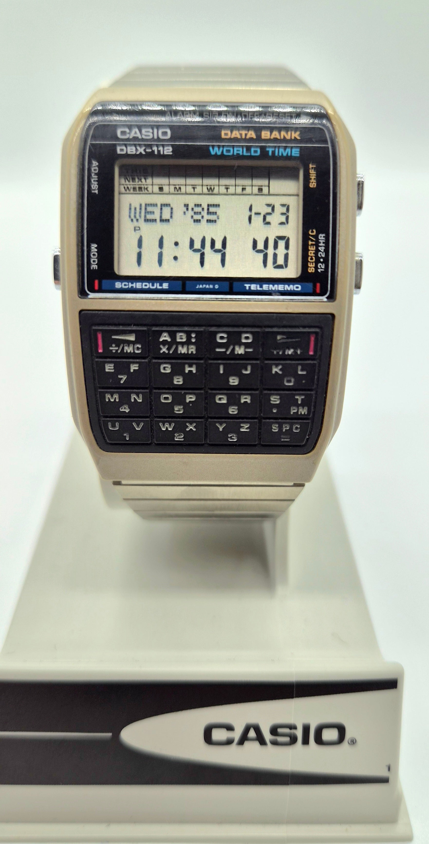 A vintage Casio DBX-118 World Time digital watch with a beige plastic case, black face, and built-in calculator keypad, displayed on a white stand.