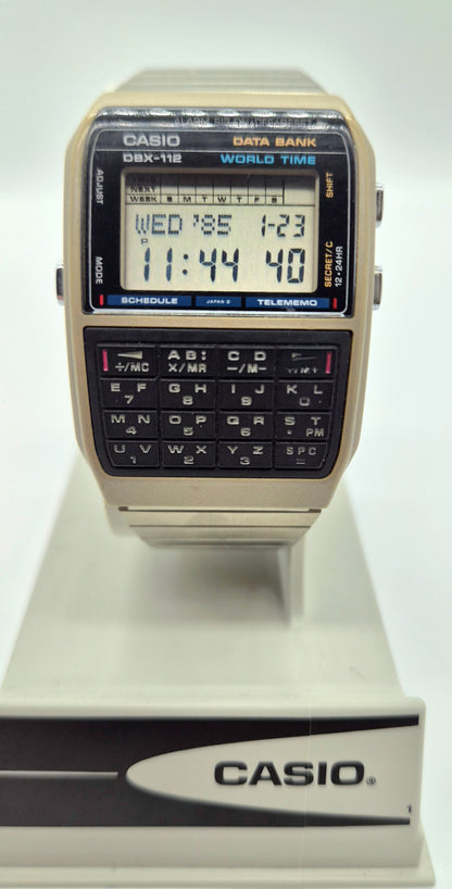 A vintage Casio DBX-118 World Time digital watch with a beige plastic case, black face, and built-in calculator keypad, displayed on a white stand.