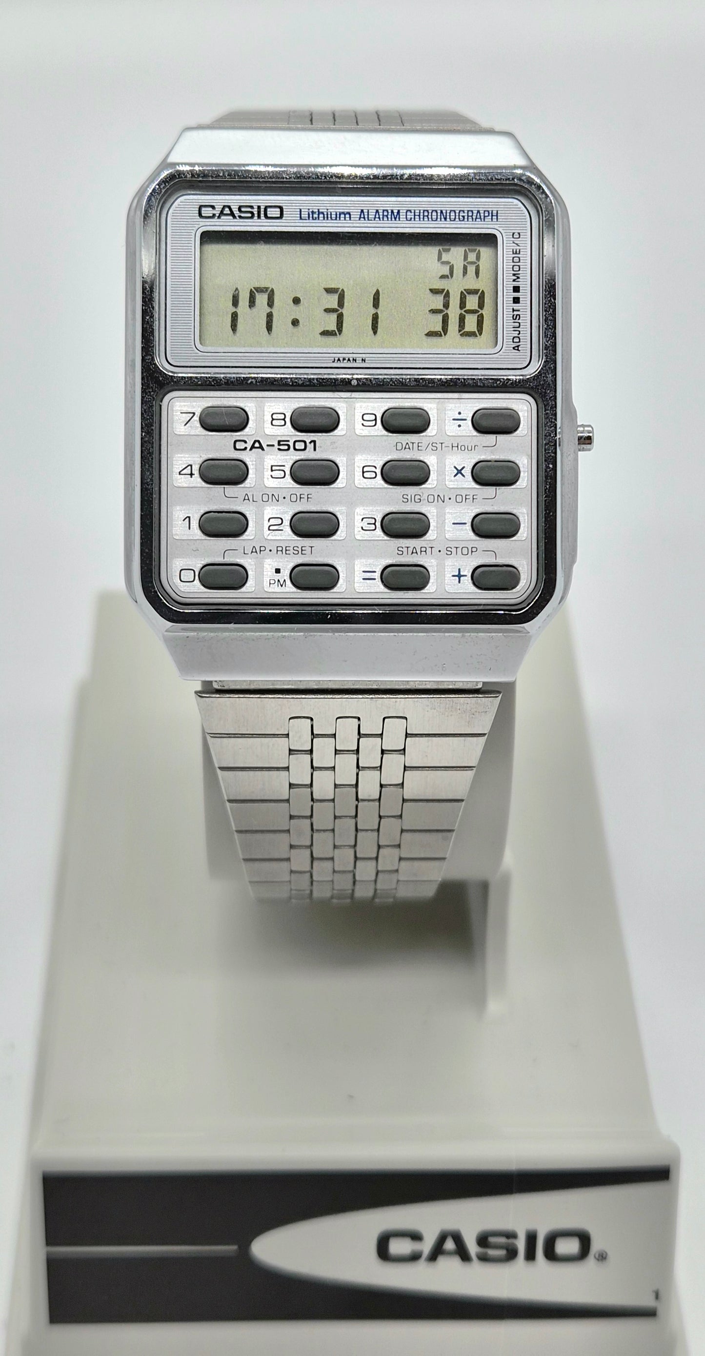 A Casio CA-S501 wristwatch with a silver metal case and link bracelet, featuring a digital display and calculator-style keypad, shown on a white display stand with a Casio box.