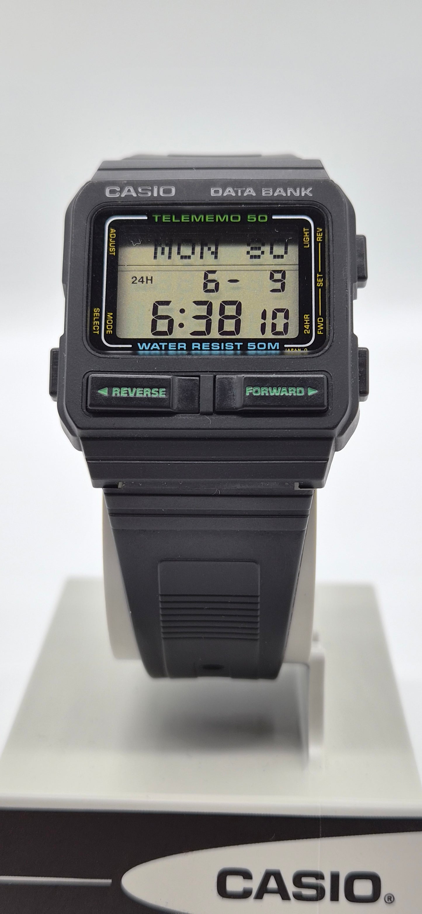 A Casio Data Bank Telememo 50 digital wrist watch with a black plastic case and strap, featuring a green and yellow LCD display, shown on a white display stand with a Casio logo box at the bottom.