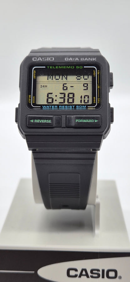 A Casio Data Bank Telememo 50 digital wrist watch with a black plastic case and strap, featuring a green and yellow LCD display, shown on a white display stand with a Casio logo box at the bottom.