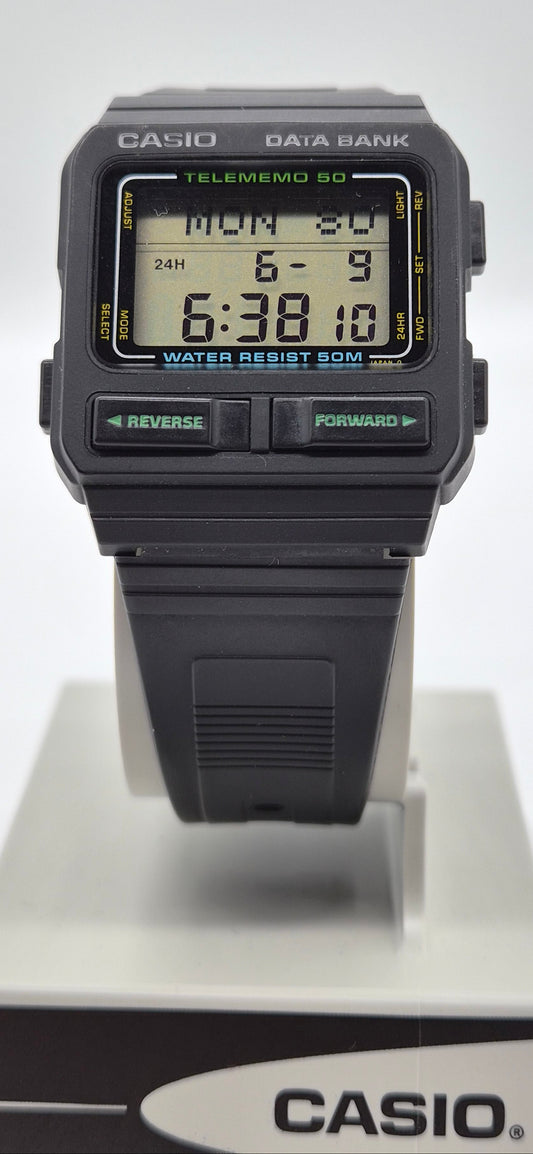 A Casio Data Bank Telememo 50 digital wrist watch with a black plastic case and strap, featuring a green and yellow LCD display, shown on a white display stand with a Casio logo box at the bottom.