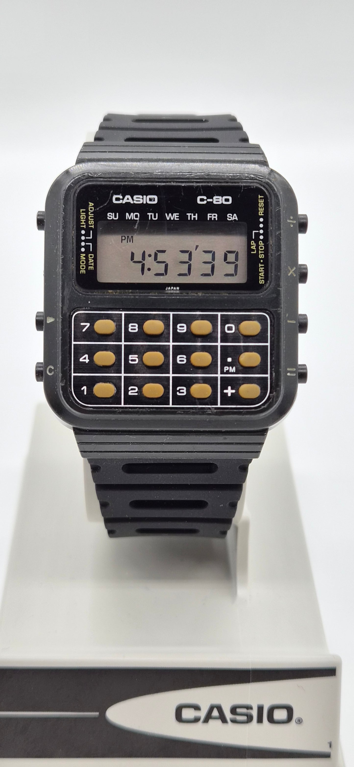 A Casio C-80 calculator watch with a black plastic case and strap, featuring gold-toned calculator buttons and a digital display, shown on a white display stand with the Casio logo.