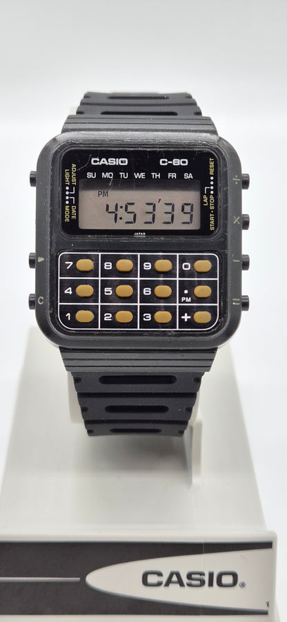 A Casio C-80 calculator watch with a black plastic case and strap, featuring gold-toned calculator buttons and a digital display, shown on a white display stand with the Casio logo.