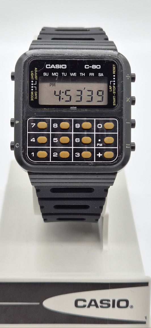 A Casio C-80 calculator watch with a black plastic case and strap, featuring gold-toned calculator buttons and a digital display, shown on a white display stand with the Casio logo.