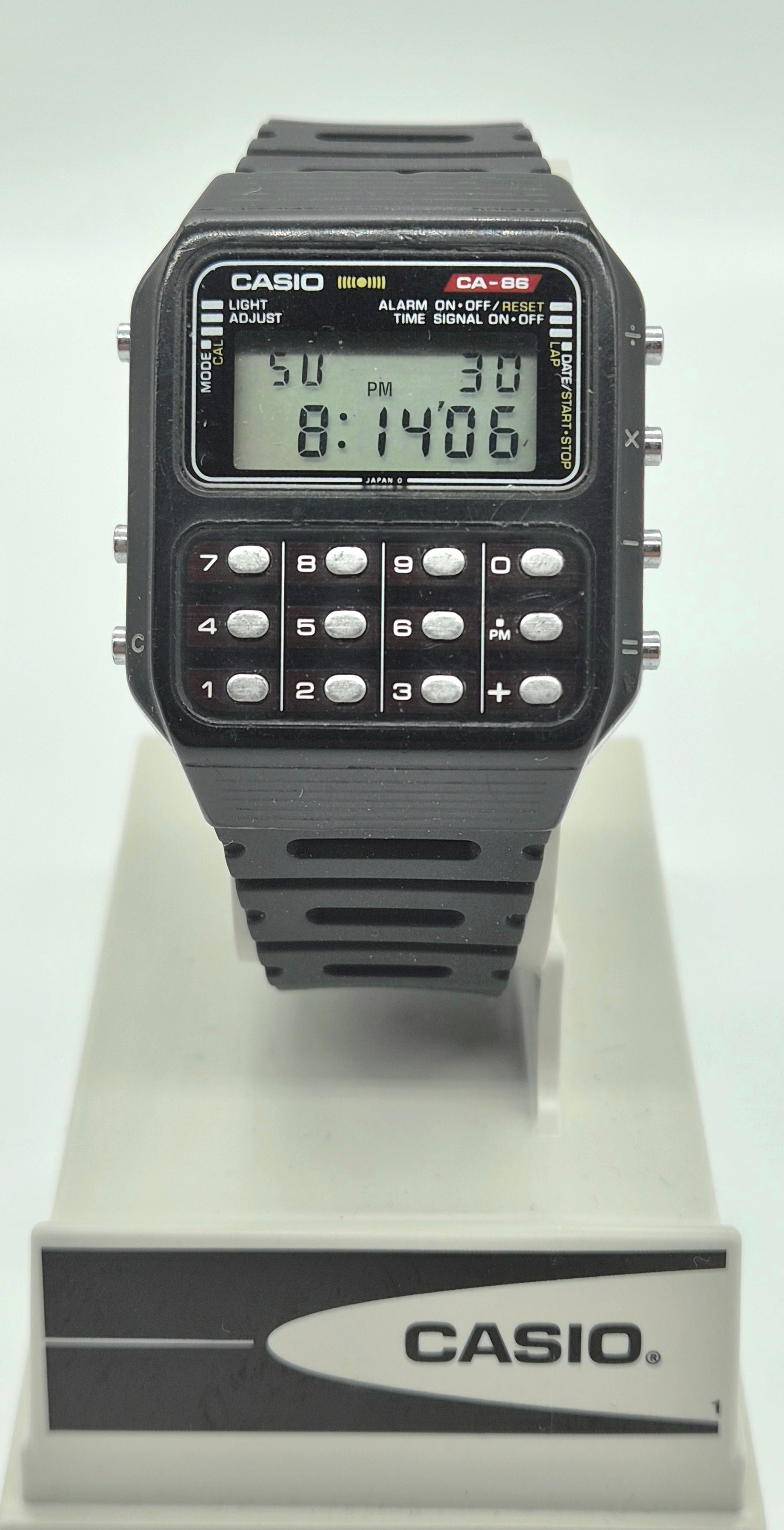 A Casio calculator-style wristwatch with a black rectangular face, digital display, and numeric keypad, mounted on a white display stand with a Casio logo.