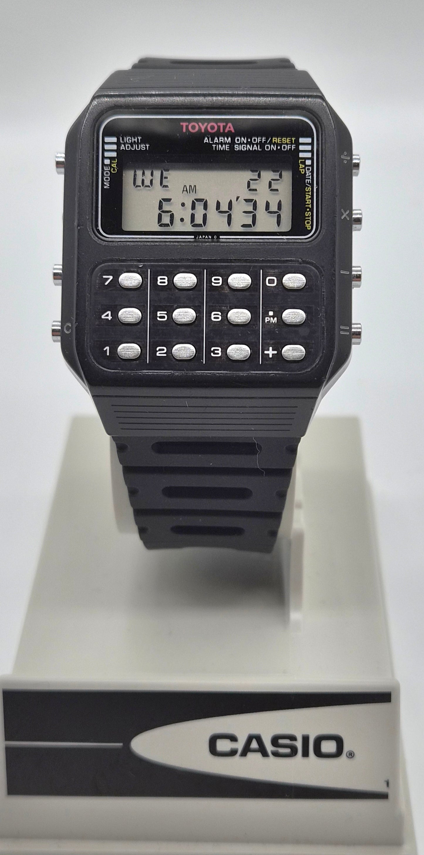 A black Casio Toymaster calculator watch with a digital LCD display and numeric keypad, mounted on a white display stand featuring the Casio logo.