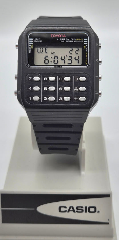 A black Casio Toymaster calculator watch with a digital LCD display and numeric keypad, mounted on a white display stand featuring the Casio logo.