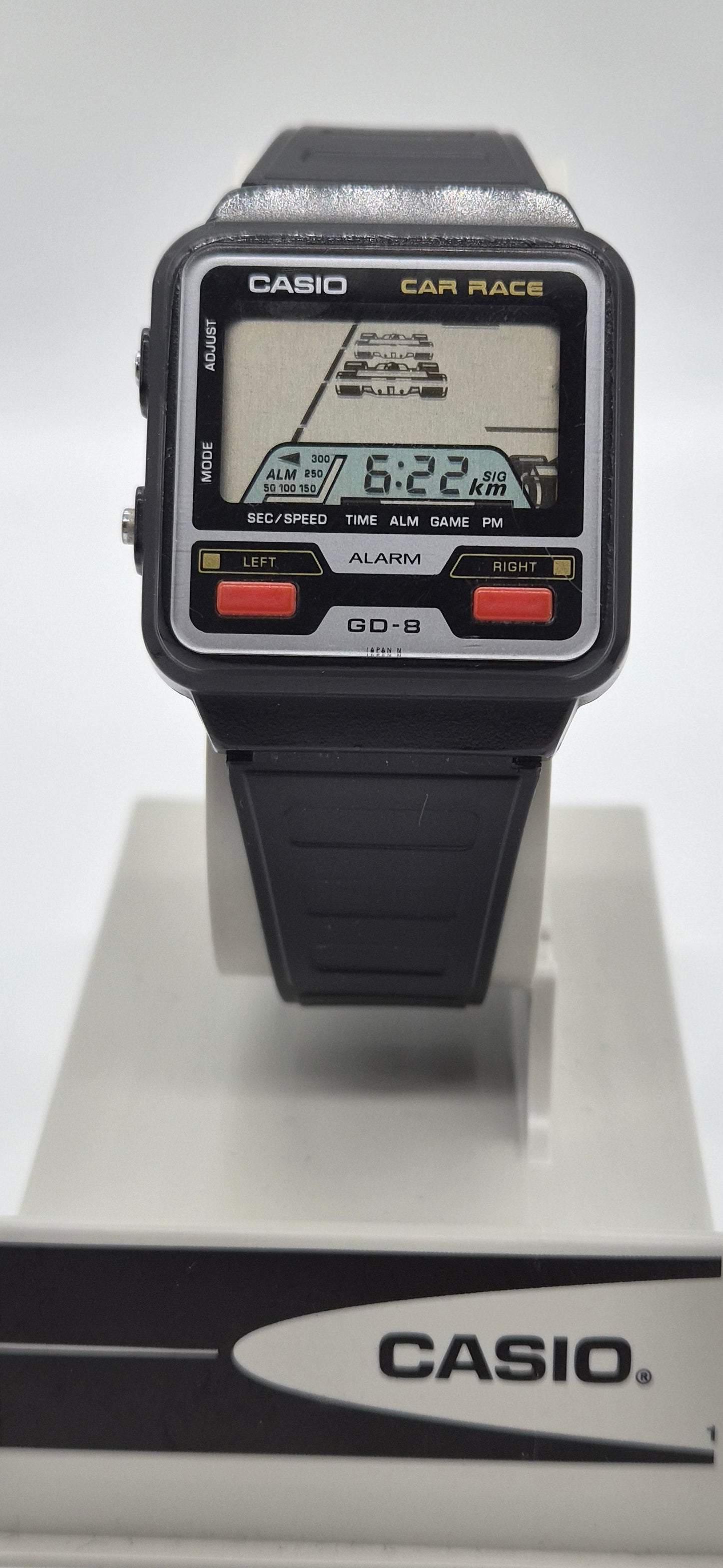 A vintage Casio car race watch with a black plastic strap and case, featuring a digital display showing a race track and the time 6:22. The watch is displayed on a white stand with a black and white Casio logo base.