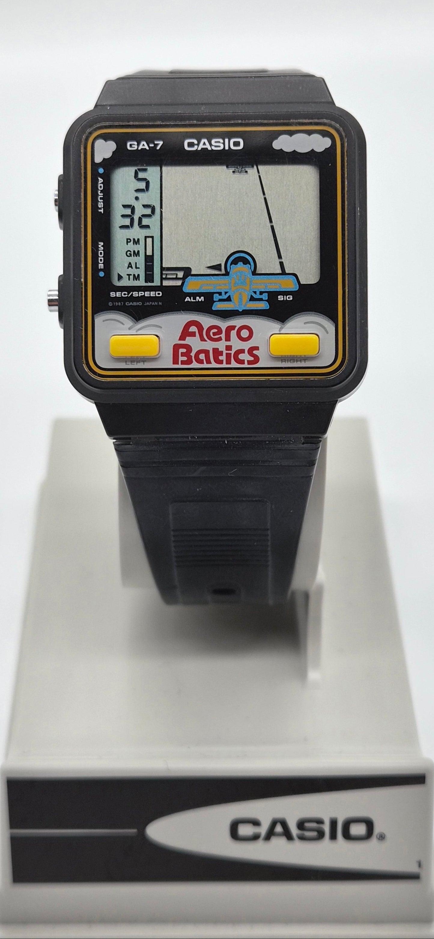 A vintage Casio GA-7 Aero Batic watch with a black plastic case and strap, featuring a colorful screen with yellow buttons, displayed on a white stand.