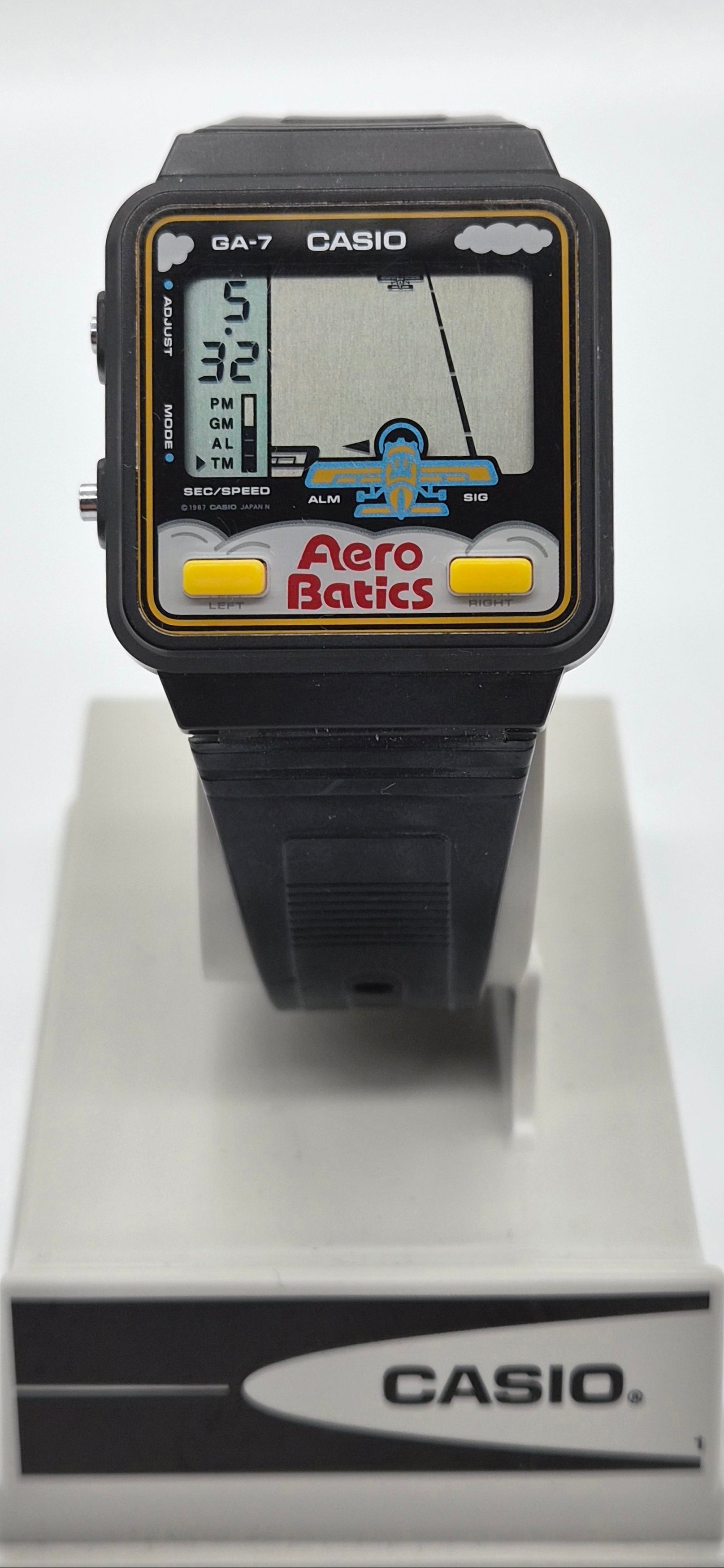 A vintage Casio GA-7 Aero Batic watch with a black plastic case and strap, featuring a colorful screen with yellow buttons, displayed on a white stand.
