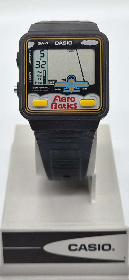 A vintage Casio GA-7 Aero Batic watch with a black plastic case and strap, featuring a colorful screen with yellow buttons, displayed on a white stand.
