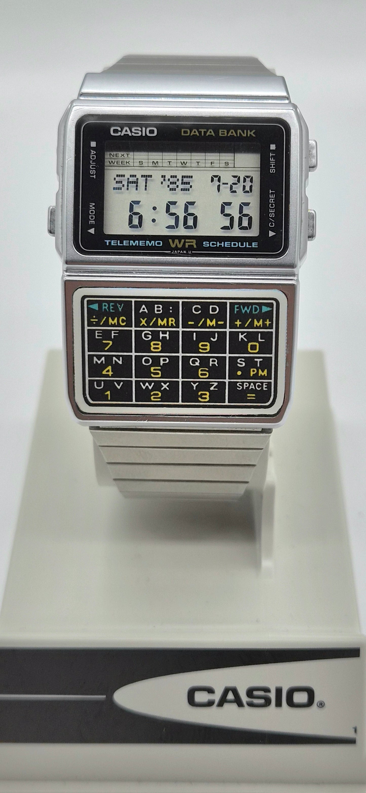 A vintage Casio Data Bank calculator watch with a silver-tone metal case, black face, and a built-in calculator keypad on the lower half, displayed on a white Casio stand against a light gray background.