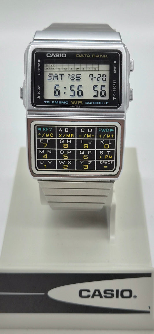 A vintage Casio Data Bank calculator watch with a silver-tone metal case, black face, and a built-in calculator keypad on the lower half, displayed on a white Casio stand against a light gray background.