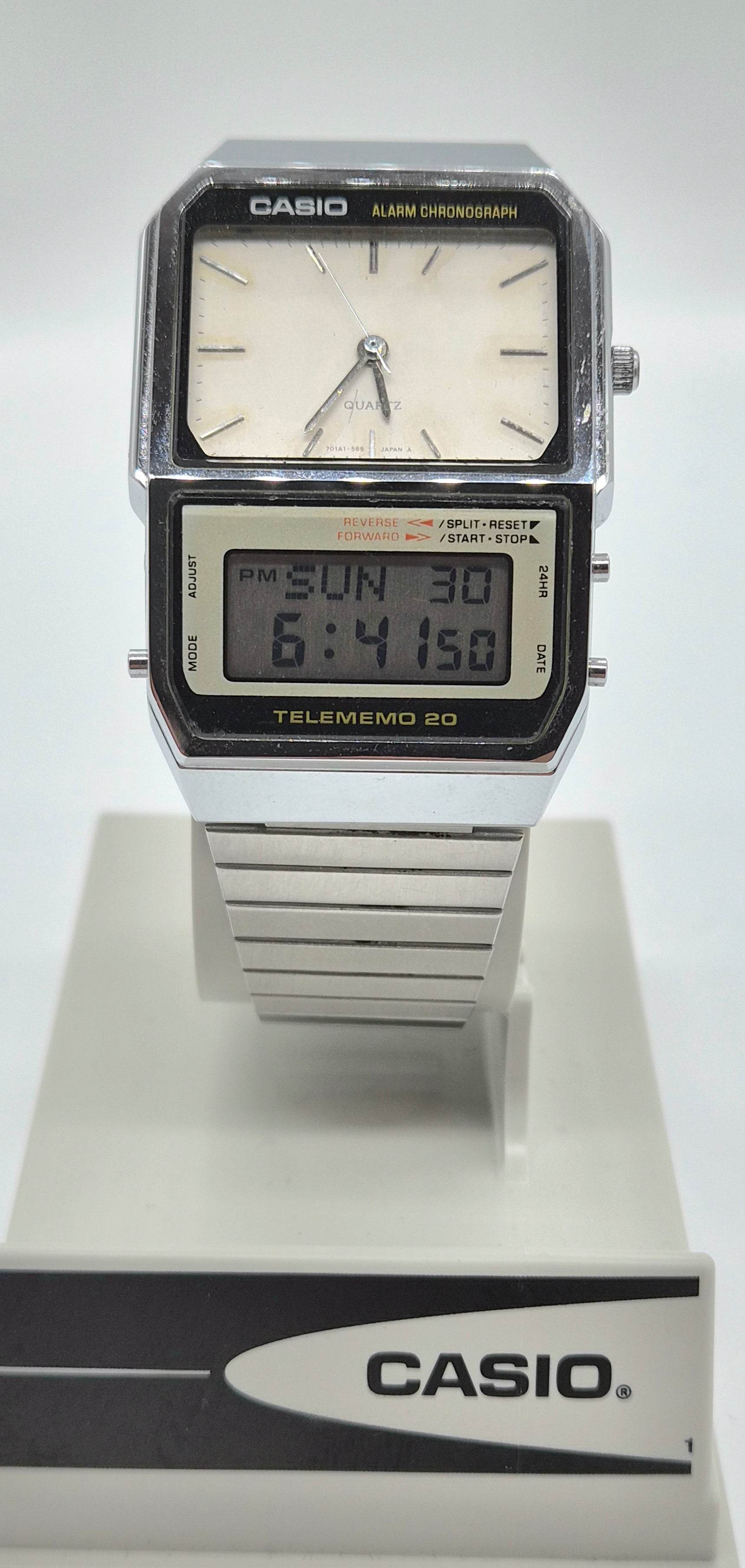 A Casio Telememo 20 digital alarm clock watch with a silver-tone metal case and strap, featuring a white analog dial and a black LCD display, shown on a white display stand with a Casio logo.