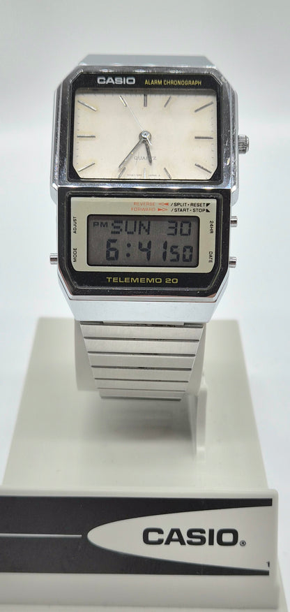 A Casio Telememo 20 digital alarm clock watch with a silver-tone metal case and strap, featuring a white analog dial and a black LCD display, shown on a white display stand with a Casio logo.