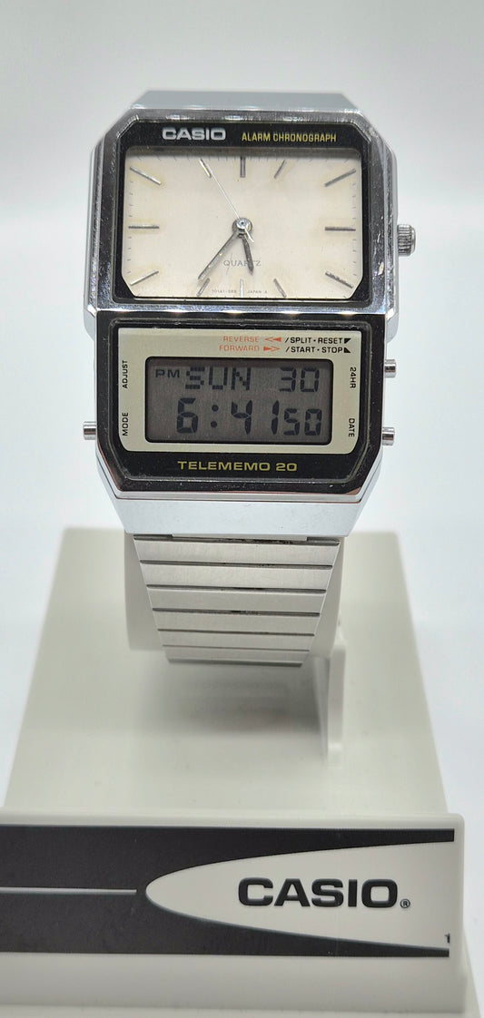 A Casio Telememo 20 digital alarm clock watch with a silver-tone metal case and strap, featuring a white analog dial and a black LCD display, shown on a white display stand with a Casio logo.