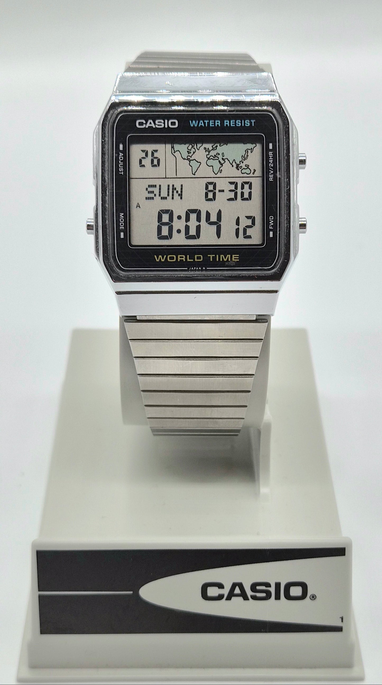 A Casio digital wrist watch with a silver-tone metal case and strap, displayed on a white stand with a black and white Casio logo plaque. The watch face is black with a digital display showing the time as 8:04 and world time settings.