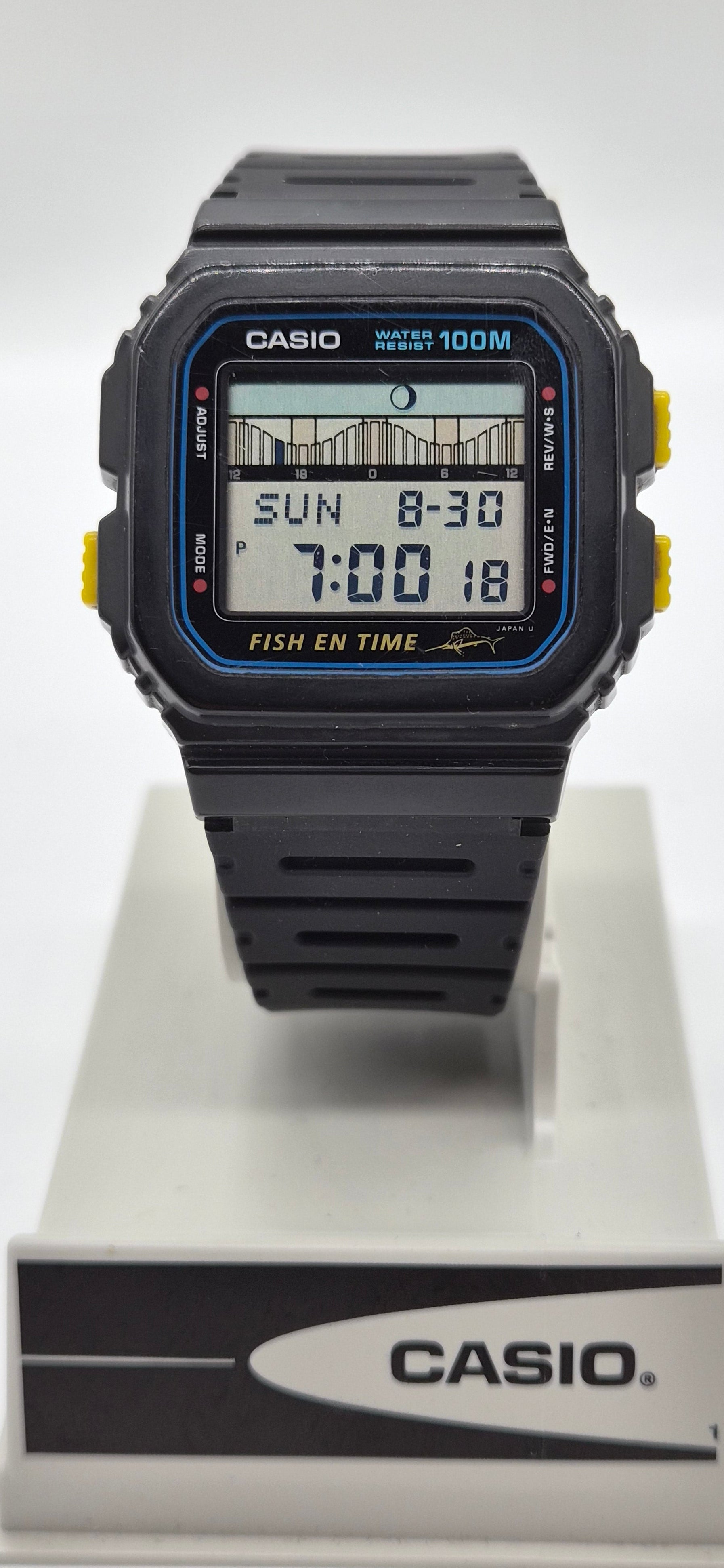 A Casio digital watch with a black resin case and strap, featuring a blue bezel accent and yellow side buttons, displayed on a white stand with a branded base.