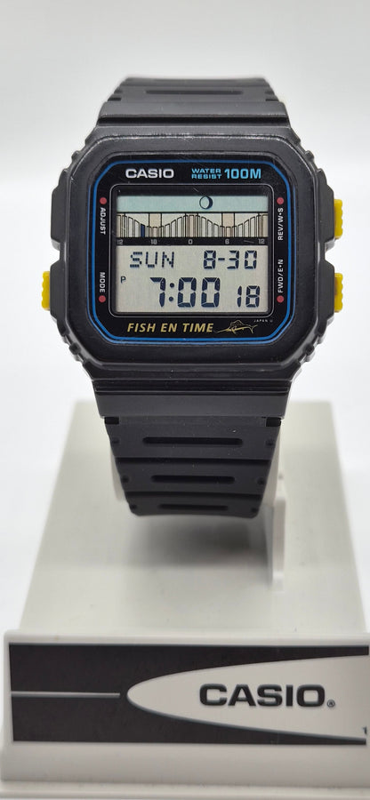 A Casio digital watch with a black resin case and strap, featuring a blue bezel accent and yellow side buttons, displayed on a white stand with a branded base.