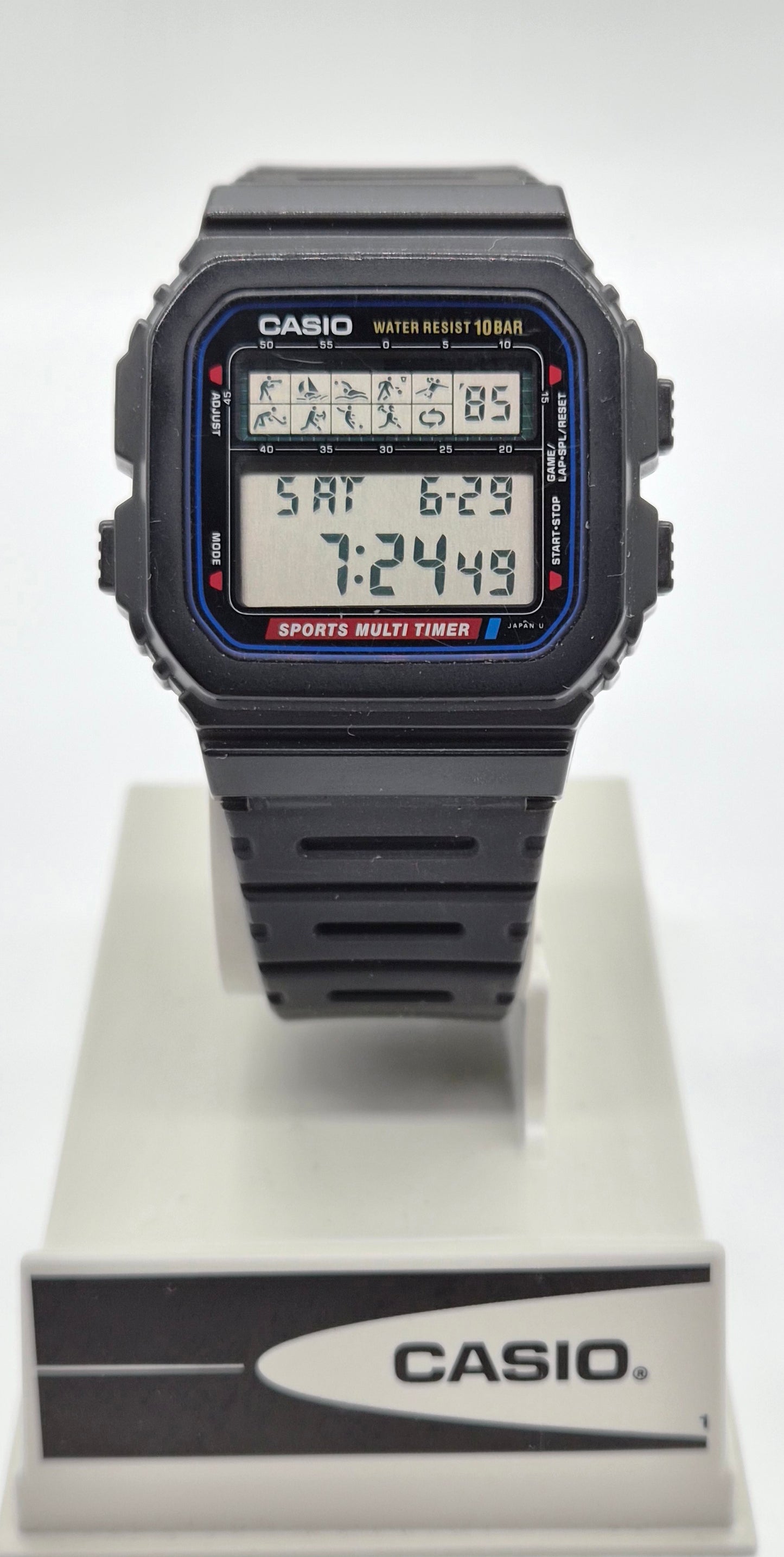A Casio digital watch with a black rectangular case and strap, featuring a blue and red bezel detail and a multi-function display, shown on a white display stand with a Casio logo.