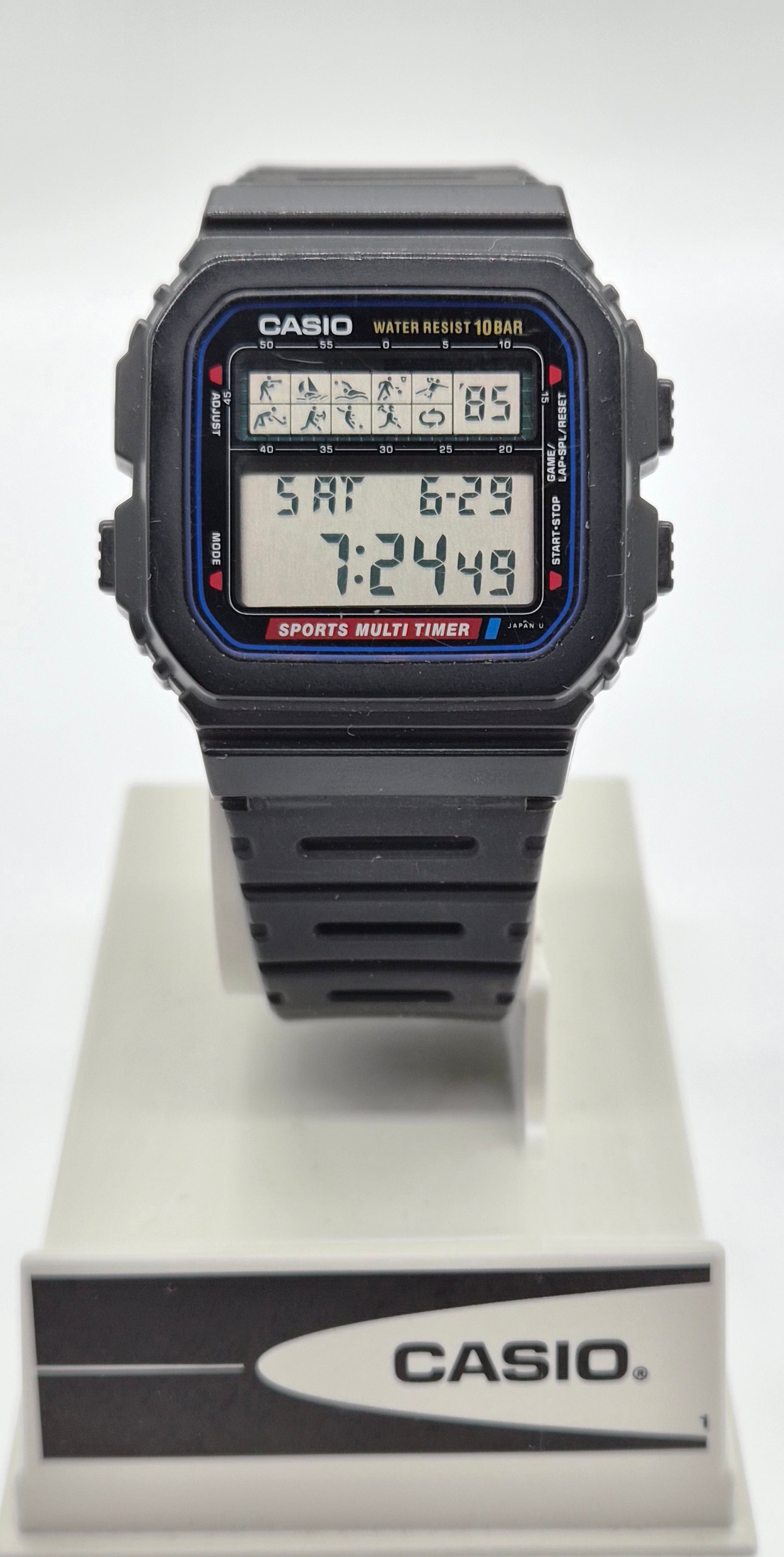 A Casio digital watch with a black rectangular case and strap, featuring a blue and red bezel detail and a multi-function display, shown on a white display stand with a Casio logo.