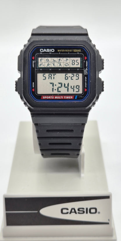A Casio digital watch with a black rectangular case and strap, featuring a blue and red bezel detail and a multi-function display, shown on a white display stand with a Casio logo.