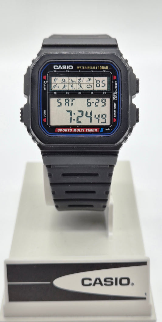 A Casio digital watch with a black rectangular case and strap, featuring a blue and red bezel detail and a multi-function display, shown on a white display stand with a Casio logo.