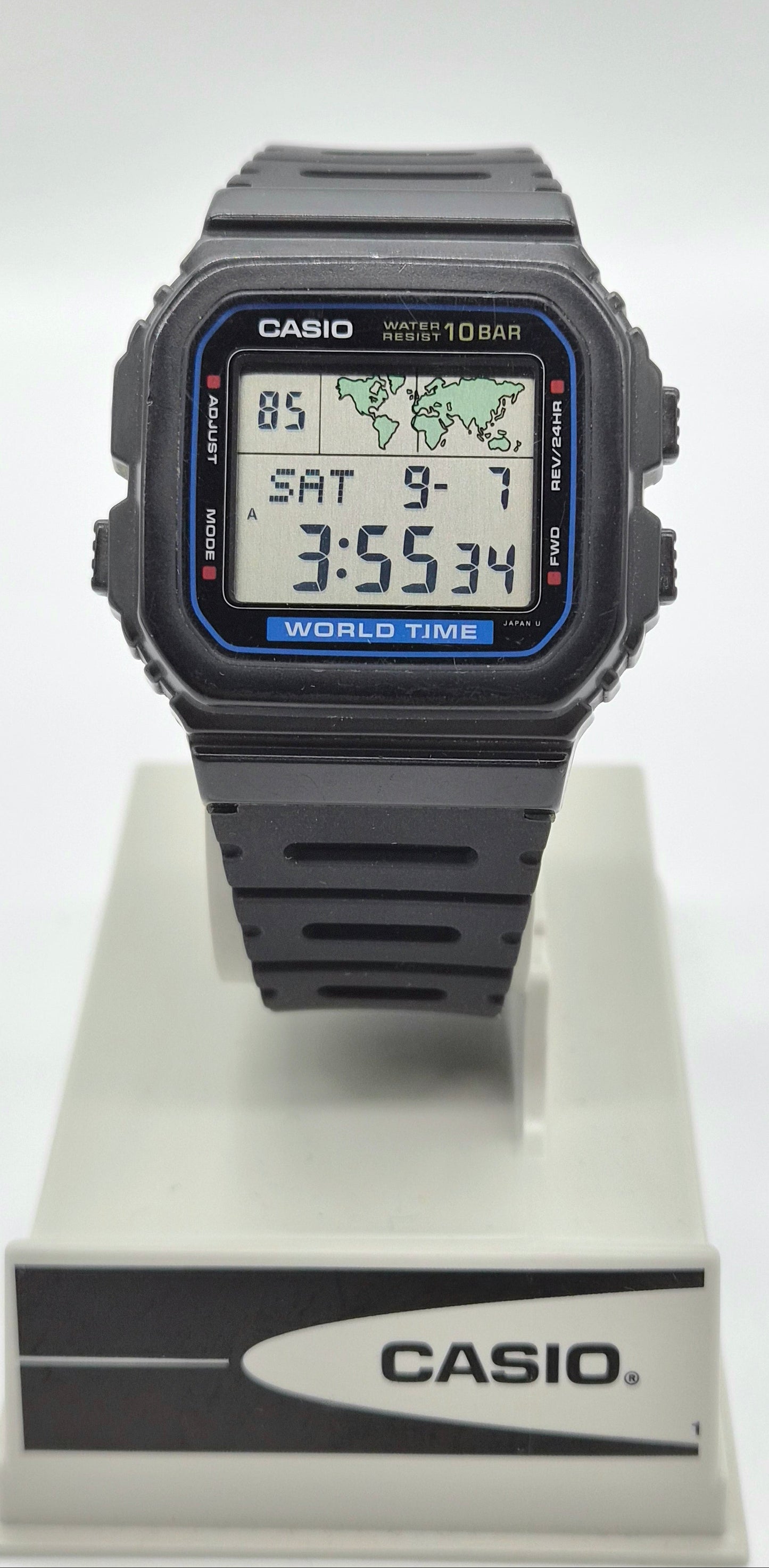 A Casio digital wrist watch with a black square case and black strap, displayed on a white stand with the Casio logo. The watch face has a blue border and digital display showing the time as 3:55:34.