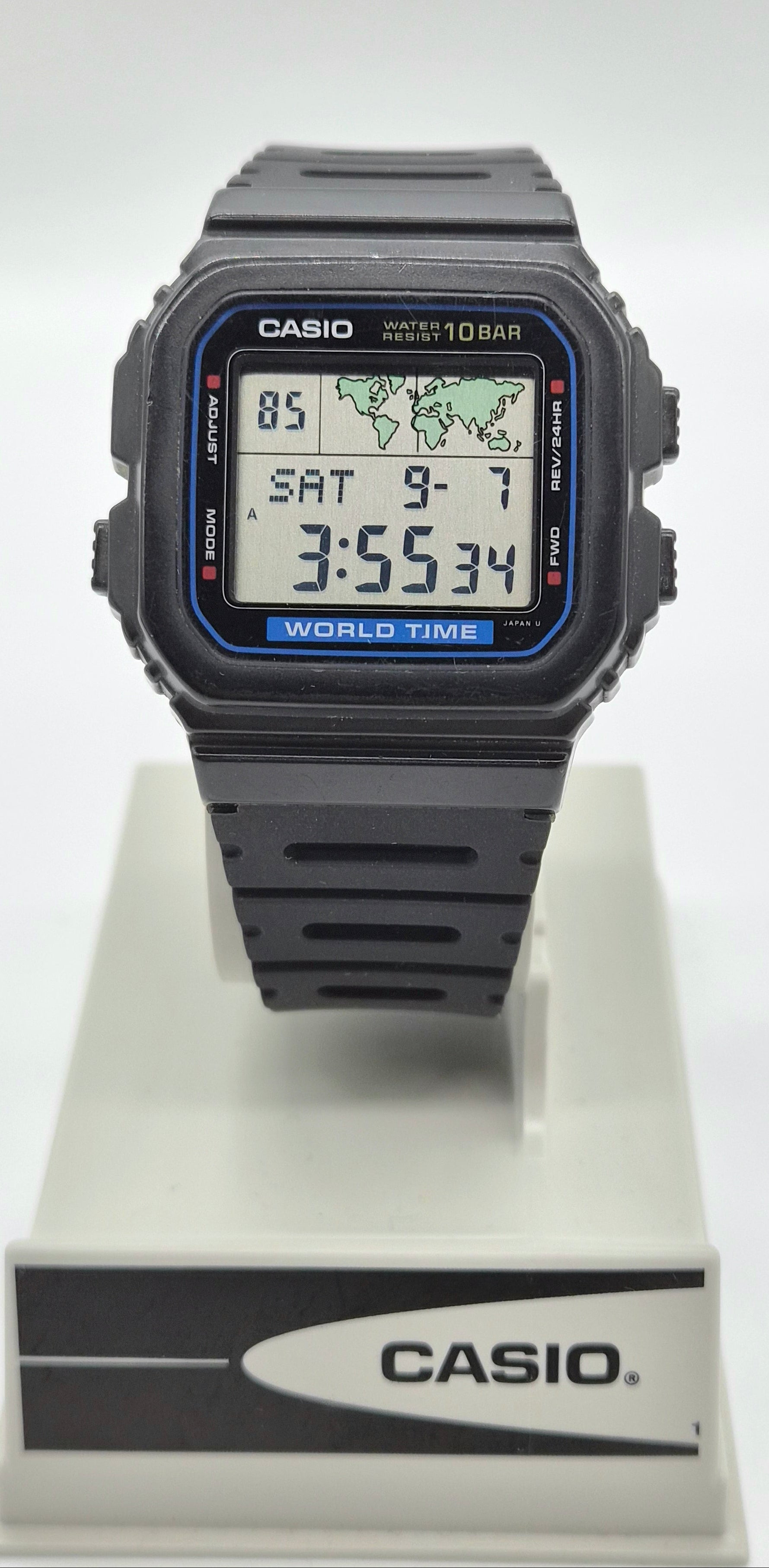 A Casio digital wrist watch with a black square case and black strap, displayed on a white stand with the Casio logo. The watch face has a blue border and digital display showing the time as 3:55:34.