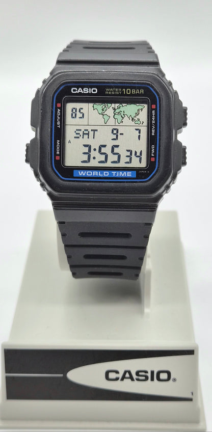 A Casio digital wrist watch with a black square case and black strap, displayed on a white stand with the Casio logo. The watch face has a blue border and digital display showing the time as 3:55:34.