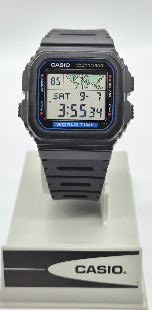 A Casio digital wrist watch with a black square case and black strap, displayed on a white stand with the Casio logo. The watch face has a blue border and digital display showing the time as 3:55:34.
