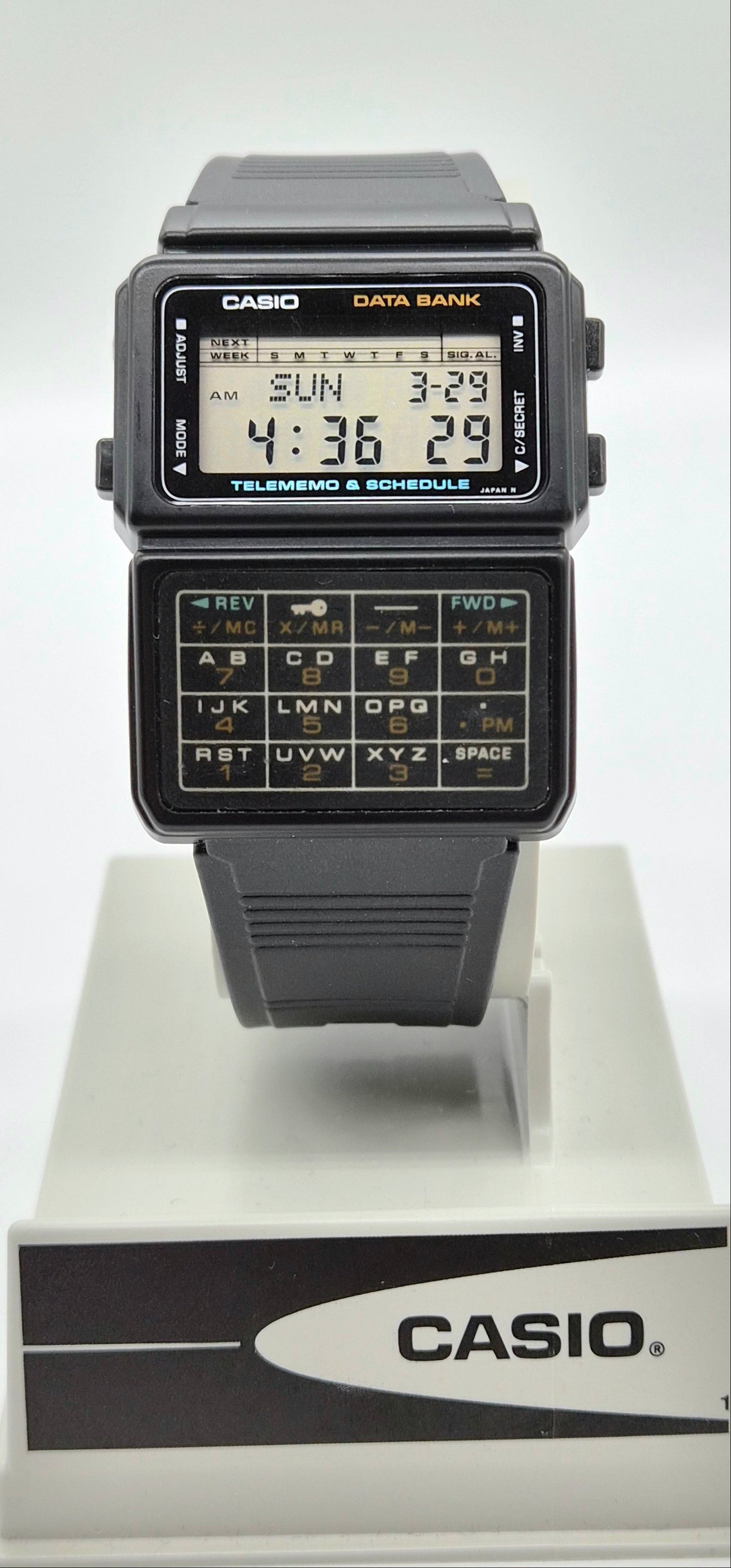 A Casio digital wristwatch with a black strap and case, featuring a built-in calculator keypad on the front. The watch is displayed on a white stand with a Casio-branded base against a plain light background.