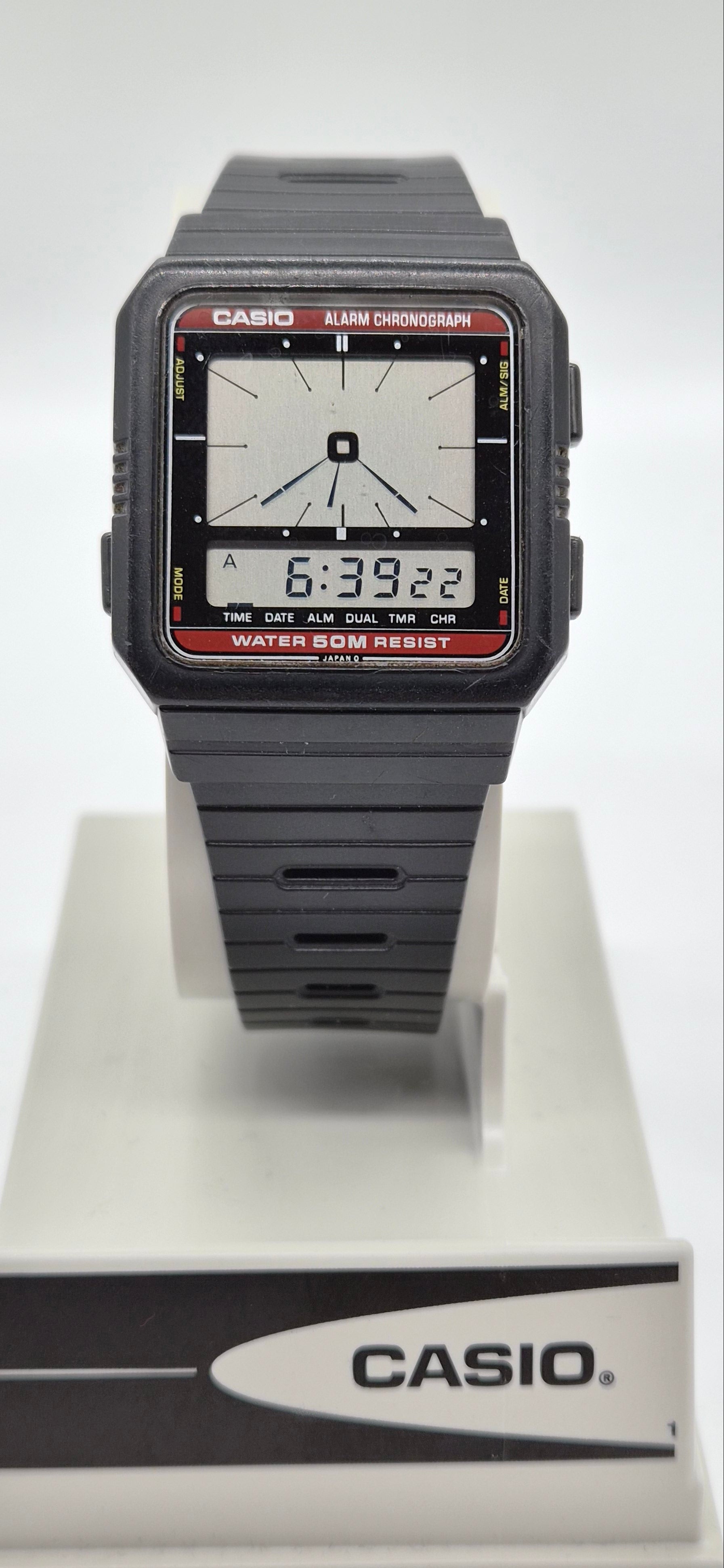 A Casio digital wristwatch with a dark gray resin case and strap, featuring a white analog display face with black hands and red bezel details, resting on a white and gray Casio display stand.