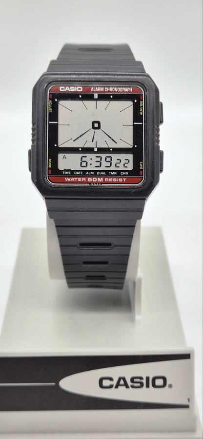 A Casio digital wristwatch with a dark gray resin case and strap, featuring a white analog display face with black hands and red bezel details, resting on a white and gray Casio display stand.