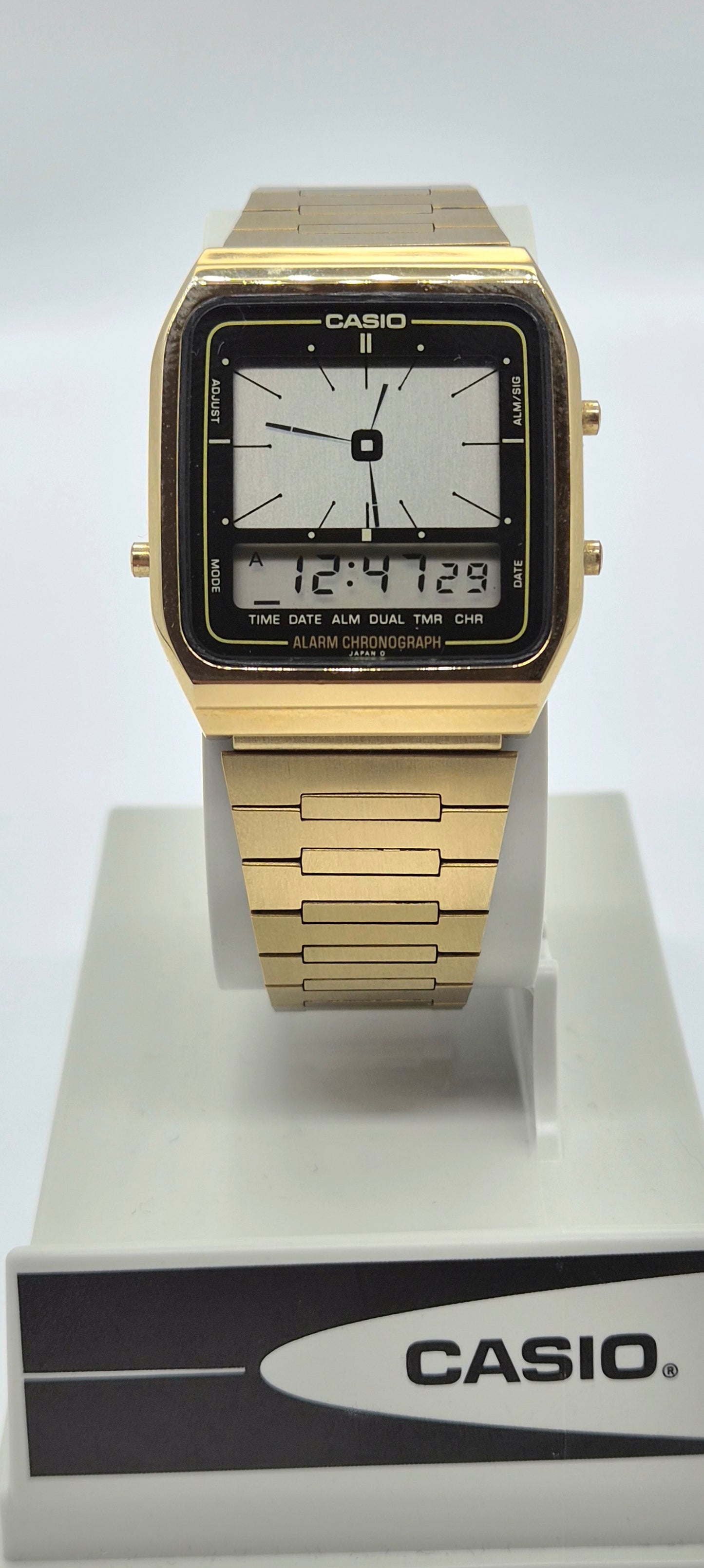 A Casio square analog watch with a gold-tone metal case and link bracelet, featuring a black bezel and a white face with a digital display overlay. The watch is displayed on a white stand with a black and white Casio logo box in the foreground.