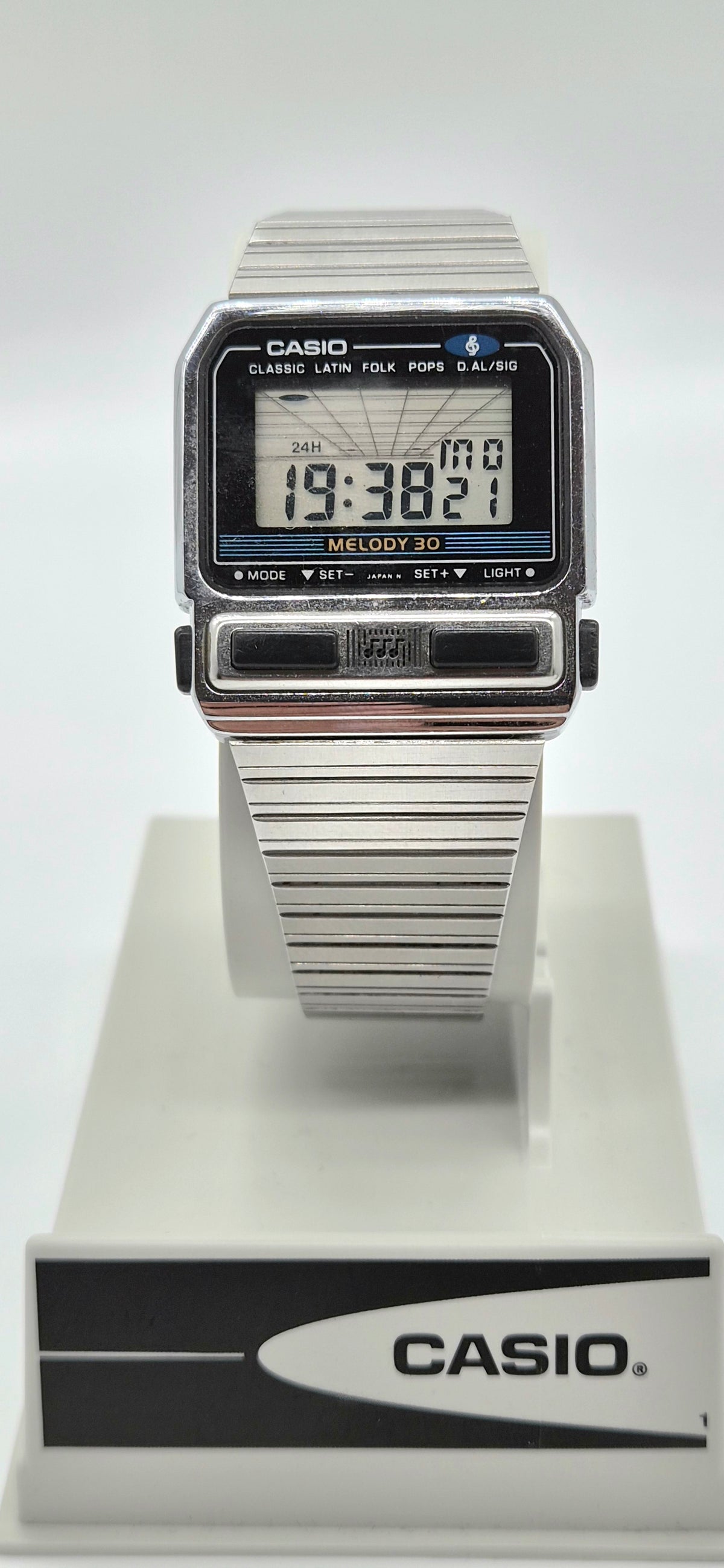 A vintage Casio digital watch with a silver-tone metal link bracelet and a black rectangular face, displayed on a white stand with a branded base.