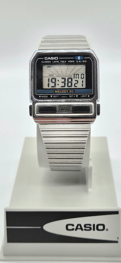 A vintage Casio digital watch with a silver-tone metal link bracelet and a black rectangular face, displayed on a white stand with a branded base.