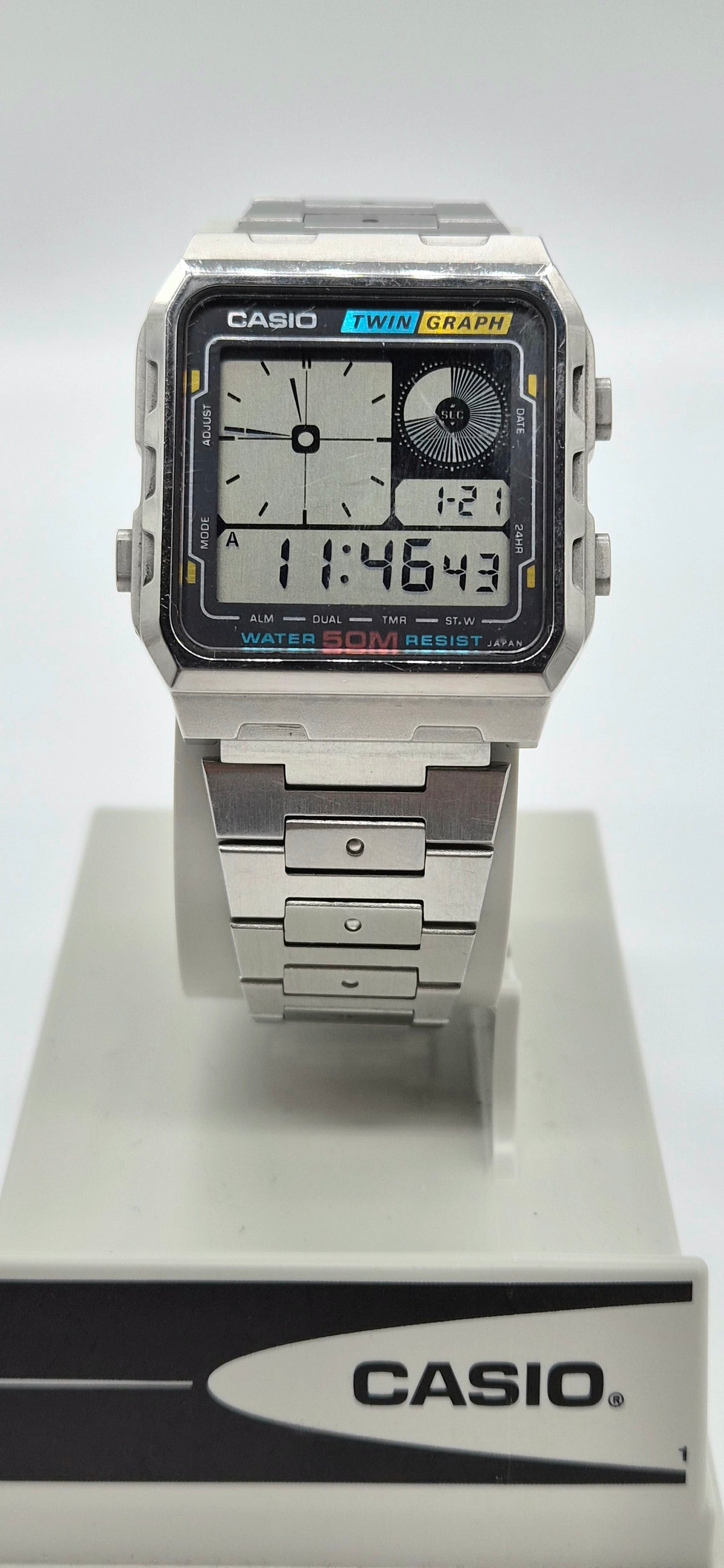A Casio dual display analog watch with a silver-toned metal case and link bracelet, mounted on a white display stand with a Casio-branded base.