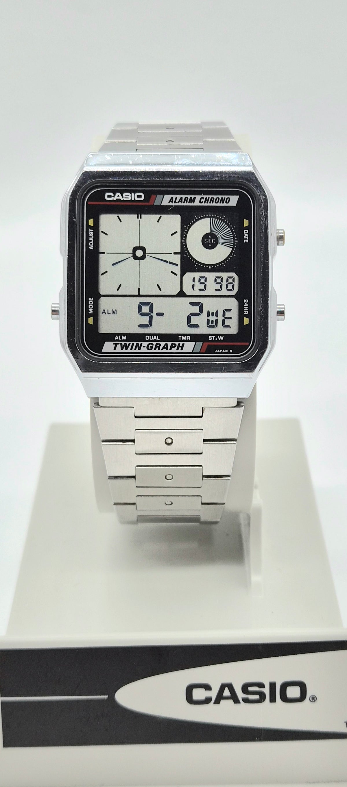 A Casio dual display analog-digital wristwatch with a silver-tone metal case and bracelet, shown standing on a white display stand with a branded base.