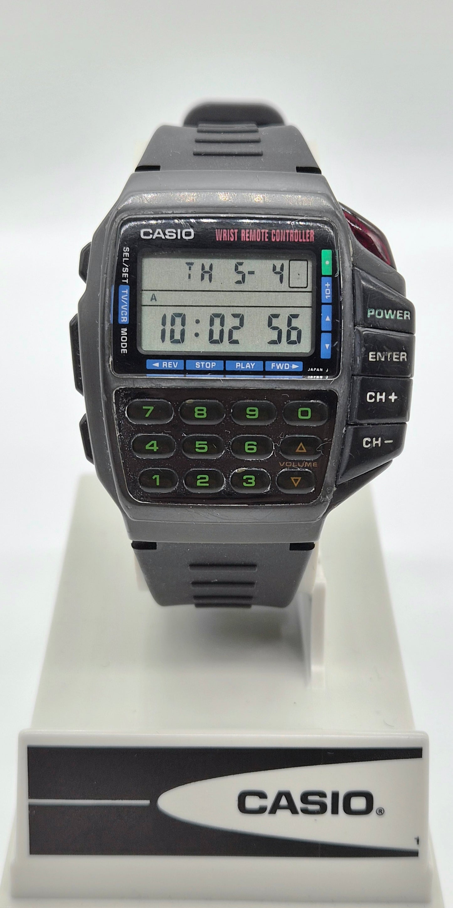 A Casio digital wrist watch with a dark gray strap and case, featuring a built-in calculator keypad and LCD display, displayed on a white stand with a Casio logo plaque.