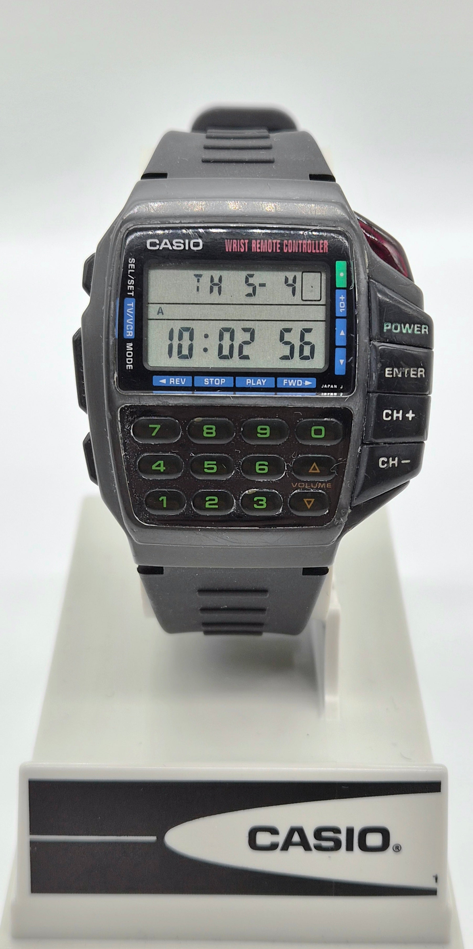A Casio digital wrist watch with a dark gray strap and case, featuring a built-in calculator keypad and LCD display, displayed on a white stand with a Casio logo plaque.