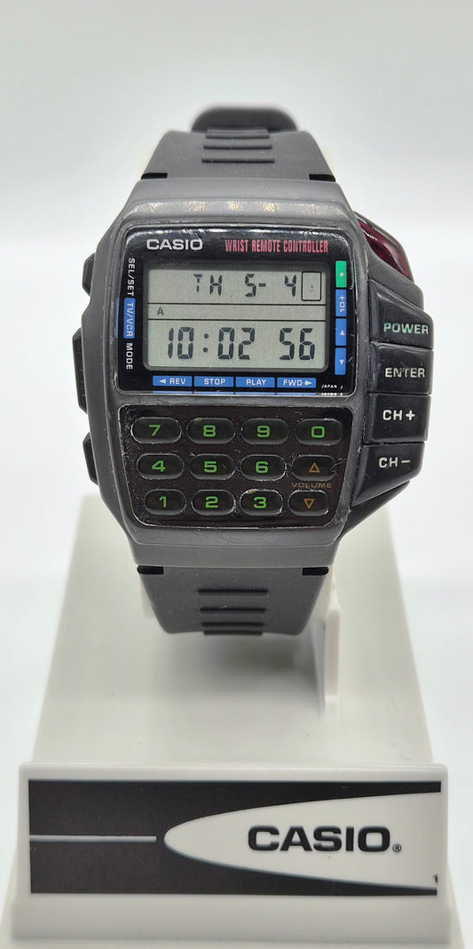 A Casio digital wrist watch with a dark gray strap and case, featuring a built-in calculator keypad and LCD display, displayed on a white stand with a Casio logo plaque.