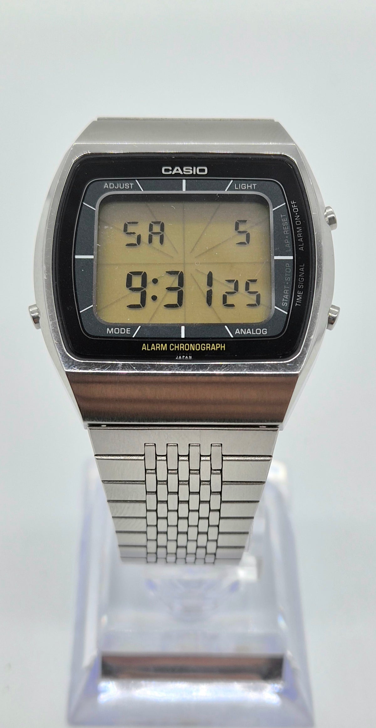 A Casio analog-digital wristwatch with a silver-tone metal case and link bracelet, featuring a black bezel and a yellow LCD display, shown on a clear plastic display stand against a white background.