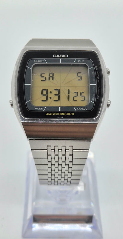 A Casio analog-digital wristwatch with a silver-tone metal case and link bracelet, featuring a black bezel and a yellow LCD display, shown on a clear plastic display stand against a white background.