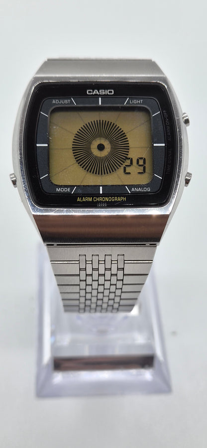 A Casio analog watch with a silver-tone metal case and link bracelet, featuring a black rectangular dial with a yellow-tinted center and digital-style numbers, displayed on a clear stand against a white background.