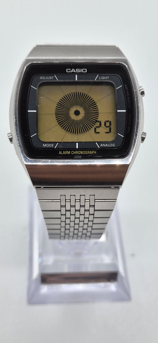 A Casio analog watch with a silver-tone metal case and link bracelet, featuring a black rectangular dial with a yellow-tinted center and digital-style numbers, displayed on a clear stand against a white background.